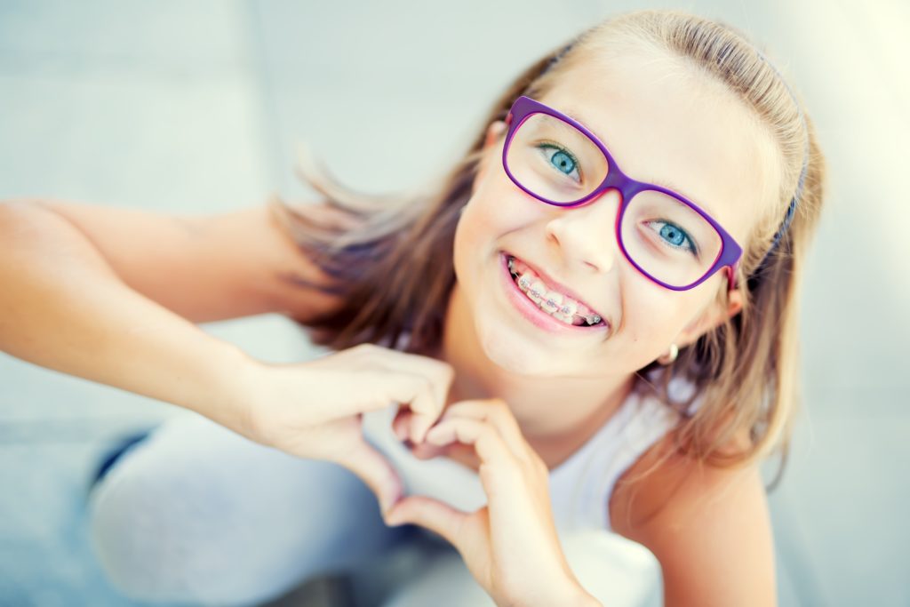Smiling little girl in with braces and glasses showing heart with hands